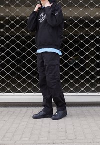 Czarna bluza z kapturem z grafiką, jasnoniebieska koszula pod spodem, czarne spodnie cargo i czarne sneakersy, stojący przed metalową siatką bramy.