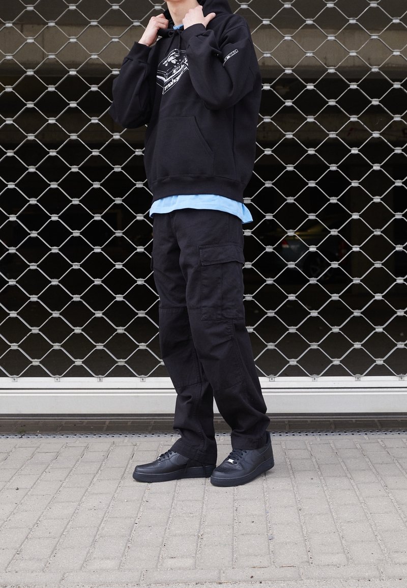 Czarna bluza z kapturem z grafiką, jasnoniebieska koszula pod spodem, czarne spodnie cargo i czarne sneakersy, stojący przed metalową siatką bramy.