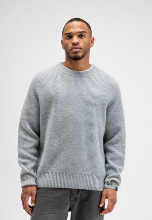 Man wearing a light gray ribbed knit sweater and dark gray pants standing against a white background.