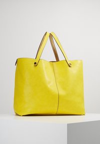 Žlutá kožená taška přes rameno se hladkým povrchem, širokým otvorem a dvěma hnědými koženými popruhy. Obsahuje jemný šev podél středu.