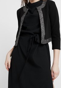 Kvinna klädd i en svart klänning med knytband i midjan och en kort svart jacka med silverglitterkant.