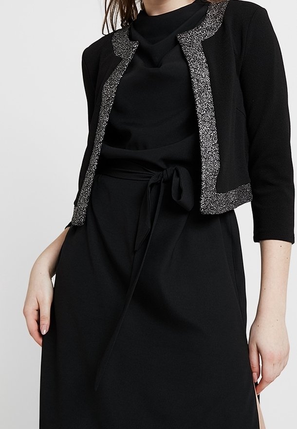 Kvinna klädd i en svart klänning med knytband i midjan och en kort svart jacka med silverglitterkant.