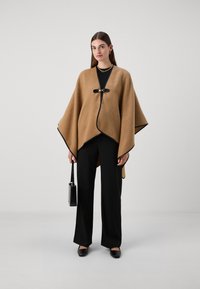 Brun uld poncho med sorte kanter og en frontlukning. Matchet med sorte bukser og en textured sort håndtaske. Glat stoftekstur.