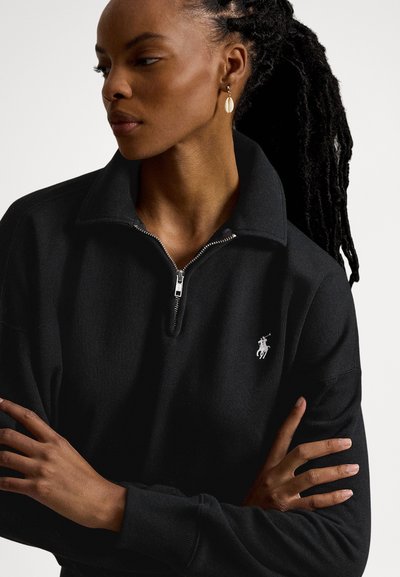 Juodas puloveris su užtraukiamu kaklu, pagamintas iš struktūrizuotos medžiagos ir su mažu baltu logotipu kairėje krūtinės pusėje. Modelis su pintais plaukais.