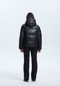 Chaqueta de plumas negra con capucha, diseño acolchonado, textura brillante y relleno grueso; combinada con pantalones negros con puños con cremallera.