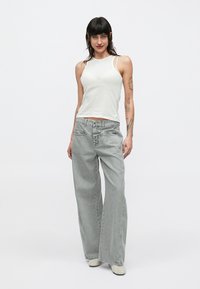 Femme aux cheveux foncés portant un haut blanc sans manches et un jean gris ample, debout avec une main dans la poche devant un fond uni.