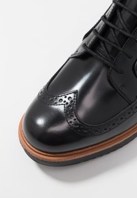 Chaussure en cuir noir avec des détails brogue, texture lisse, embout rond et semelle extérieure contrastante couleur tan. Équipée de lacets noirs et d'œillets.