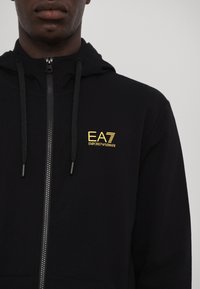 Zwarte zip-up hoodie met een gouden EA7 Emporio Armani-logo op de linkerkant van de borst, met trekkoorden en een zachte textuur.