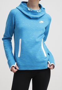 Blå Nike-tröja med vita dragkedjefickor och tumhål, bärs av person med händerna i fickorna, mot en enfärgad bakgrund.