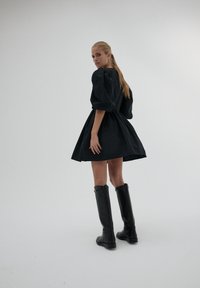 Kobieta o długich blond włosach, ubrana w krótką czarną sukienkę z bufiastymi rękawami oraz czarne skórzane buty do kolan, stojąca na jednolitym tle.