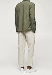 Giacca camicia verde oliva con vestibilità rilassata, abbinata a pantaloni tapered beige chiaro e sneaker bianche. Tessuto liscio e design minimale.