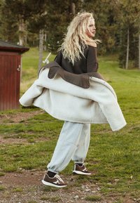 Giacca beige foderata in sherpa, pantaloni sportivi grigi e sneakers marroni con accenti crema. Il modello è in piedi sull'erba, circondato da alberi.