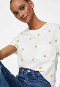 Camiseta blanca de manga corta con estampado de sol amarillo y un bolsillo en el pecho. Combinada con jeans de cintura alta azules. Textura suave.