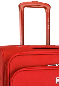 Maleta roja con ruedas, fabricada en un tejido texturizado, con cremallera superior y un asa extensible que presenta ranuras verticales. Logo de la marca visible en el asa.