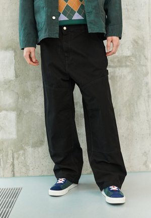 Personne portant un pantalon noir à jambes larges, des chaussures de tennis colorées, une veste verte et un pull à motifs, debout sur un sol bleu clair contre un mur en béton.