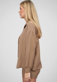 Camicia oversize marrone, con texture, maniche lunghe e chiusura a bottoni, indossata con pantaloni corti coordinati, mostrata di profilo su uno sfondo neutro.