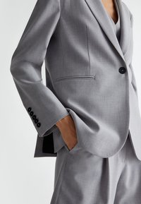 Blazer grigio su misura in tessuto testurizzato con rever a punta, chiusura a un bottone e dettagli con quattro bottoni ai polsini.