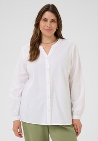 Femme avec de longs cheveux bruns, portant un chemisier blanc à boutons et un pantalon vert, se tenant contre un fond clair. 