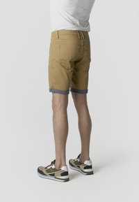 Férfi felhajtott khaki rövidnadrágban, fehér pólóban és bézs, zöld, fekete sportcipőben, háttal áll a kamerának.