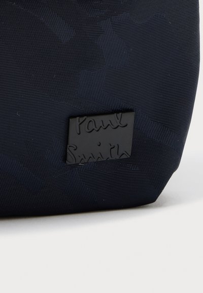 Σκούρο μπλε υφασμάτινο με υφή, με μια μαύρη ορθογώνια ετικέτα που έχει ανάγλυφη την επιγραφή "Paul Smith" με καλλιγραφικά γράμματα.
