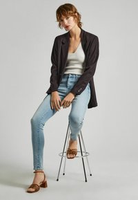 Blazer oscuro sobre una camiseta de tirantes clara y canalé, combinado con jeans skinny azul claro y sandalias de tacón con tiras marrones. Fondo liso.