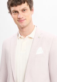 Helle rosa Blazer mit Revers, strukturiertem Stoff und einem weißen Taschentuch. Getragen über einem cremefarbenen Poloshirt mit Kragen. Silberner Reversaufsatz sichtbar.