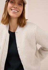 Veste bomber en polaire blanche avec un tissu texturé et un col côtelé gris, superposée sur un haut sombre, présentée sur un fond neutre.