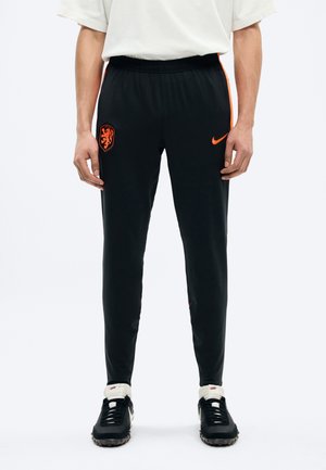 Personne portant un pantalon de sport Nike noir avec un emblème de lion orange et le logo Nike, assorti à des baskets Nike noires et blanches.