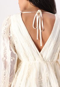 Abito bianco in pizzo con profondo scollo a V sul retro, maniche lunghe trasparenti e chiusura con nastro al collo, arricchito da dettagli di ricamo floreale.