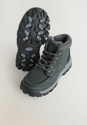 Bottes en cuir vert foncé avec des accents gris, une semelle en caoutchouc texturée et des lacets gris. Elles présentent un col rembourré et une étiquette de logo sur le côté.