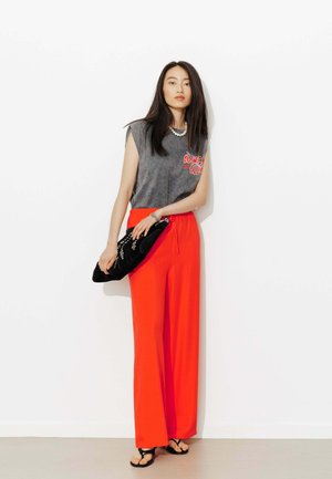 Femme debout contre un mur blanc, portant un top sans manches gris, un pantalon large rouge vif, des sandales noires et tenant une pochette noire.