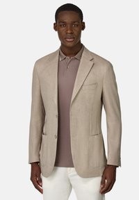 Blazer de tejido beige con dos bolsillos frontales, solapas con muesca y dos botones. Lleva puesto encima de una camisa polo marrón, que presenta una textura suave.