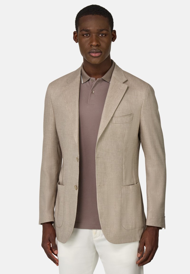 Blazer de tejido beige con dos bolsillos frontales, solapas con muesca y dos botones. Lleva puesto encima de una camisa polo marrón, que presenta una textura suave.