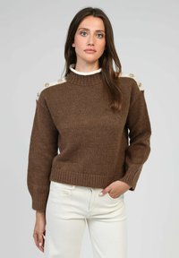 Pull marron tricoté avec un col montant, comportant des accents blancs sur les épaules et des boutons décoratifs. Ourlet côtelé et manches longues.