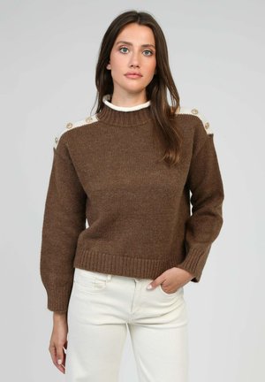 Pull marron tricoté avec un col montant, comportant des accents blancs sur les épaules et des boutons décoratifs. Ourlet côtelé et manches longues.
