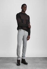 Musta pyöreäkauluksinen neulepaita, jossa on pieni logo, vaaleanharmaat slim-fit farkut ja tummanruskeat nauhalla kiinnitettävät saappaat. Sileät pinnat ja minimalistinen muotoilu.