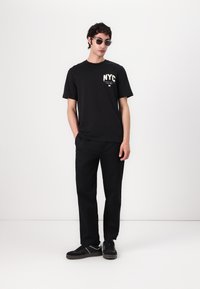 T-shirt noir à manches courtes avec un graphique "NYC" sur la poitrine gauche, associé à un pantalon noir et des baskets noires, porté avec des lunettes de soleil grises.