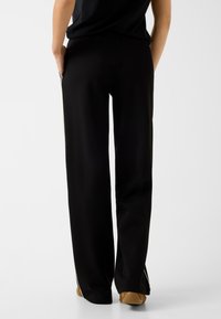 Person, die schwarze Hosen mit weitem Bein und Seitenstreifen sowie beige Sneakers trägt, steht mit den Händen in den Taschen, von hinten gesehen.