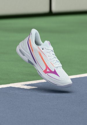 Chaussure de sport blanche avec des accents violets et orange flottant au-dessus d'un court de tennis près de la ligne de démarcation bleue et verte.