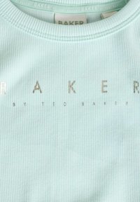 Ljusblått ribbat tyg med metallisk silvertext som visar "BAKER" och "by TED BAKER" i ett modernt typsnitt. Rund halsringning.