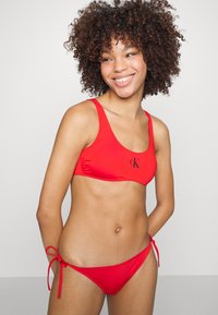Rød bikiniset laget av glatts materiale. Toppen har rund hals og logo, mens bunnen har knytinger i sidene. Rent design, minimal aksent.