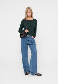 Haut à manches longues vert texturé, jean large taille haute bleu clair, et un petit sac brun foncé. Modèle portant des chaussures à enfiler bordeaux.
