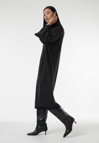 Vestido maxi negro con estampado, de mangas largas, cuello alto y tejido texturizado, acompañado de botas negras de tacón alto hasta la rodilla.