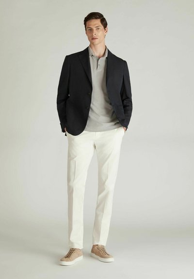 Blazer blu navy sopra un polo grigio chiaro, abbinato a pantaloni crema e sneakers beige. L'outfit mette in risalto una vestibilità su misura e texture sottili.