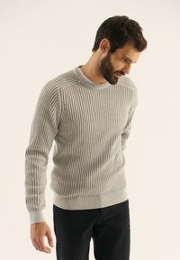 Suéter de punto acanalado gris claro con cuello redondo y mangas largas, que presenta un dobladillo ajustado y una superficie texturizada. Combinado con pantalones negros.