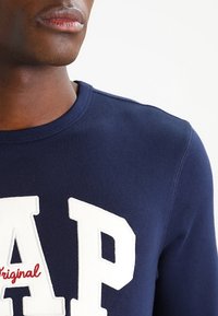 Gros plan d’une personne portant un sweat-shirt bleu marine avec de grandes lettres blanches "A P" et le mot "original" en rouge sur la poitrine.