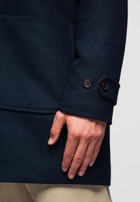 Marine uldfrakke med struktureret overflade, der har en knappet manchet og en sidelomme, holdt af en hånd mod en lys baggrund.