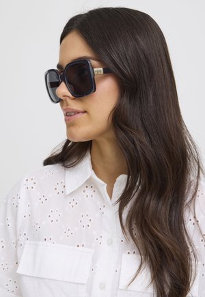 Femme aux longs cheveux noirs portant de grandes lunettes de soleil noires carrées et une chemise blanche à boutons avec œillets, regardant vers la gauche.