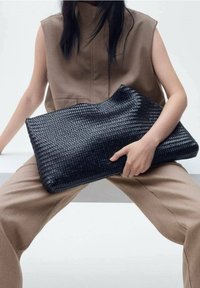 Personne dans une tenue sans manches brune tenant un grand sac à main tissé noir tout en étant assise, les jambes écartées sur un banc blanc.