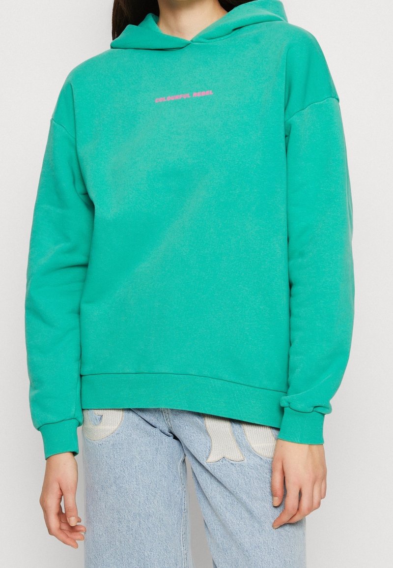 Personne portant un sweat à capuche turquoise avec un texte rose "COLOURFUL REBEL" et un jean bleu clair avec des patchs décoratifs blancs.